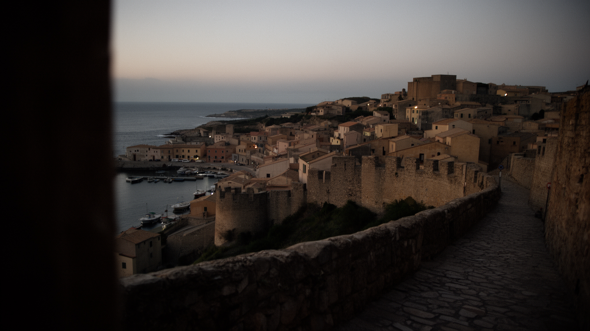 Panoràmica des del mar davant la ciutadella d'Alguer: muralla alta, teules càlides i campanar incomplert sobre una badia tranquil·la.