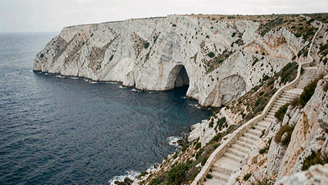 Vinyeta de la peça: Escullera alta amb arc natural a contrallum, mar plomall sota penya-segat blanc i escala llarga baixant en ziga-zaga.
