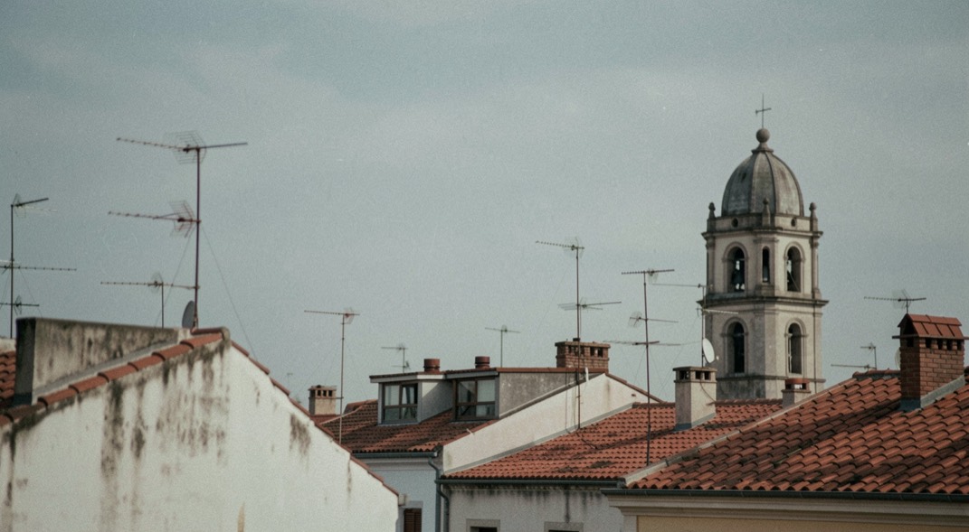 Vista horitzontal de terrats veïns amb antenes velles i un campanar incomplert al fons sota cel gris blau tranquil.